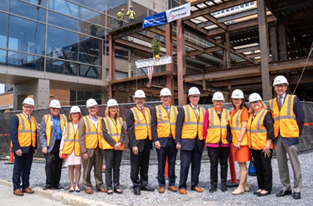 A group of “topping out” event attendees gather for a picture while the steel beam is lifted into place behind them